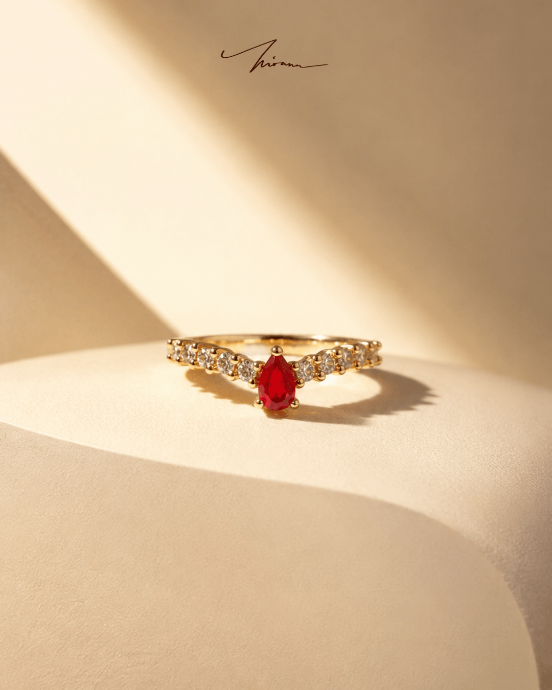 Cincin ruby pear-cut di atas suede krem dengan cahaya dramatis.